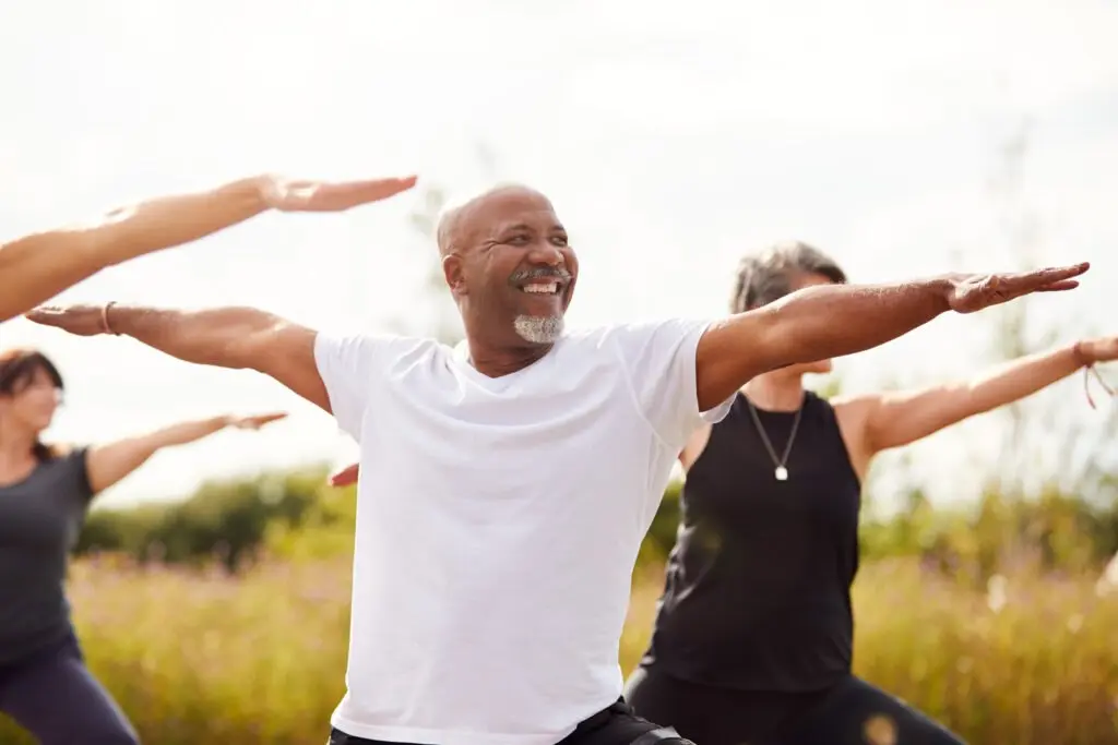 A smiling man in a white T-shirt stretching his arms wide in an outdoor group exercise class