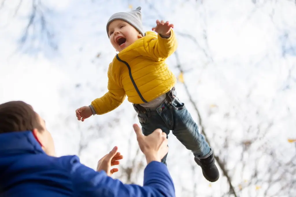 A laughing toddler in a yellow jacket tossed up in the air by a parent on a snowy day